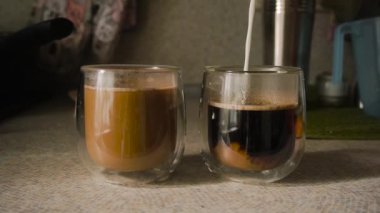 In kitchen, countertop, milk poured into two glasses, cups with double bottom with black coffee. Kitchen utensils background. Morning, bright sunbeams fall from window. Glass shade, steam. Static