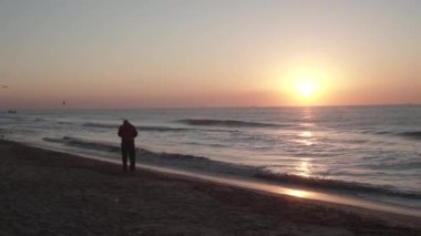 a man in a red jacket lights a cigarette and continues walking on the sand. you can see the setting sun, the birds fly low, the waves roll slowly. ships on the horizon