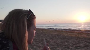 a girl with, sunglasses, blond hair smokes electronic cigarette and enjoys sea view. dawn, seagulls circling in search food, early morning, cool. On sandy beach by sea, against backdrop sunset, dawn
