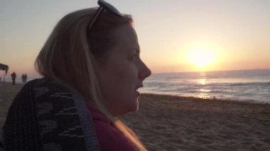 a young blonde woman with sunglasses on her head, tells stories, gesticulates while enjoying the sunrise on the black sea coast. pink sun is rising, waves are rolling, seagulls are looking for food