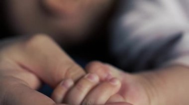 Yakın plan. Bebek küçük elleriyle annenin başparmağını sıkıca tutuyor. Yüzü bulanık çocukların arka planına karşı