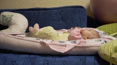 joyful little baby, lies calmly, puts hands and fingers mouth, tastes, plays alone with himself, has fun, examines room. child childrens overalls, on bed cocoon. Side view