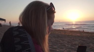 a blonde woman with sunglasses on head takes a selfie on a smartphone while enjoying the sunrise on the black sea coast. pink sun is rising, the waves are rolling, the seagulls are looking for food