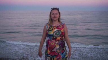 blonde in a colored dress looks directly into the camera, smiles easily, waves her hands. behind the sun sets, a warm summer breeze, the wind blows lightly, the waves roll calmly