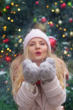 happy blond girl in winter coat and red scarf and knitted mittens blow snow close up portrai
