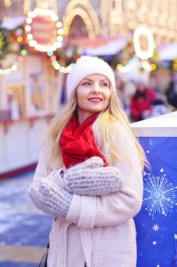 Kışlık ceketli, kırmızı atkılı ve örgü eldivenli mutlu sarışın kız karları şişiriyor. Noel arkaplanındaki portreyi kapat.