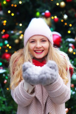 Kışlık ceketli, kırmızı atkılı ve örgü eldivenli mutlu sarışın kız karları şişiriyor. Noel arkaplanındaki portreyi kapat.