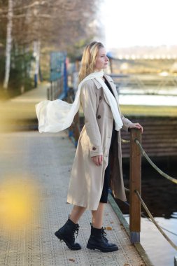 Uzun saçlı, genç, sarışın, trençkot giyen, şehir parkının arka planında sokak portresi çeken bir kadın.