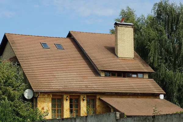 brown tiled roof of a private house with small windows and one brick chimney in green vegetation against a blue sky