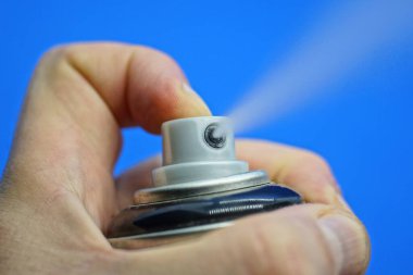 Bir el siyah bir deodorant tutuyor ve bir parmak mavi arkaplanda beyaz bir sprey ile gri düğmeye basıyor.