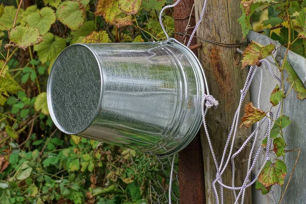 一只灰色的金属锌桶挂在街道上的绿色植被中 井边有一根白色绳子的木杆上 — 图库照片