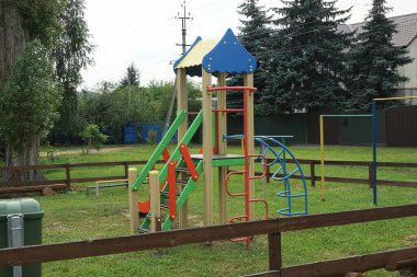Kahverengi ahşap bir çitin arkasındaki yeşil çimlerin içinde oyun parkında merdiveni olan renkli ahşap bir ev.