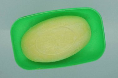 Sarı bir kalıp sabun, gri bir masada açık yeşil bir plastik sabun kabında yatıyor.