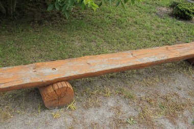 Parktaki yeşil çimlerin yanında gri zeminde duran büyük kahverengi ahşap bir bank.