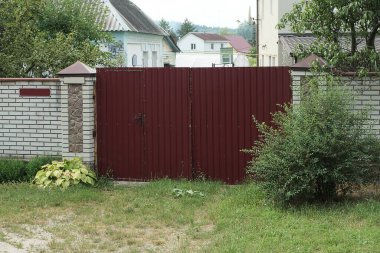 Kapalı kırmızı metal bir kapı ve yeşil bitki örtüsü ve sokakta çimen olan beyaz bir duvar.