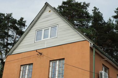 Beyaz plastik tavan arası ve gri gökyüzüne ve yeşil çam arka planına bakan camları olan kahverengi beton duvarın bir kısmı.