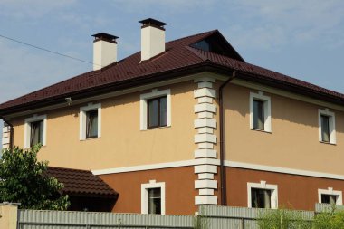 Kahverengi kiremit çatılı pencereleri olan, gri bir çitin arkasında mavi gökyüzüne karşı bacaları olan büyük, özel bir ev.