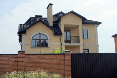 Gökyüzüne karşı tuğla bir çitin arkasında pencereleri ve açık demir bir balkonu olan büyük kahverengi özel bir ev.