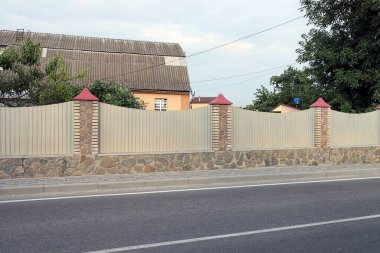 Asfalt yolun kenarındaki bir sokakta gri metal ve kahverengi taşlardan yapılmış uzun bir çit.