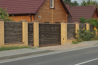 Kahverengi ahşap bir kapı ve gri asfalt bir yolun yakınındaki kırsal bir sokakta tuğla ve kalaslardan yapılmış uzun bir duvar.