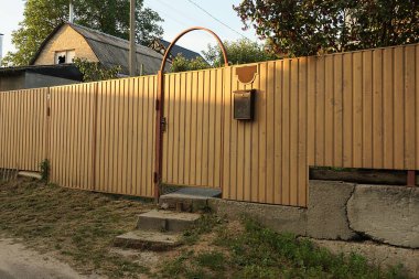 Kırsal bir sokakta, gri beton bir temelde, demir çit duvarında kapalı kahverengi metal bir kapı.