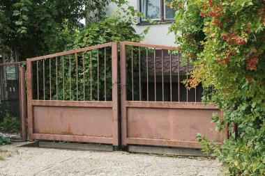 Kırsal bir sokakta, yeşil bir bitki örtüsünün üzerinde paslanmış eski bir demir kapı.