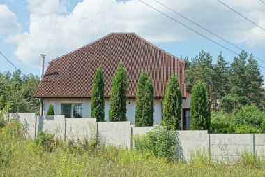 Kahverengi kiremitli bir çatı altında özel bir ev ve kırsal bir sokakta yeşil çimenli bir çitin gri beton duvarının arkasında bir sıra dekoratif kozalaklı ev.