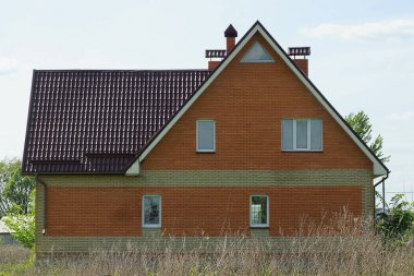 Çitin arkasında pencereleri olan, gökyüzü ve bulutların arka planında kuru gri çimenler olan kahverengi, kırmızı bir tuğla evin ön cephesi.