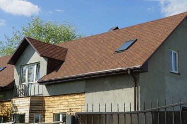 Mavi gökyüzüne karşı kahverengi kiremitli çatının altında pencereleri ve demir bir balkonu olan gri beton bir evin ön cephesi