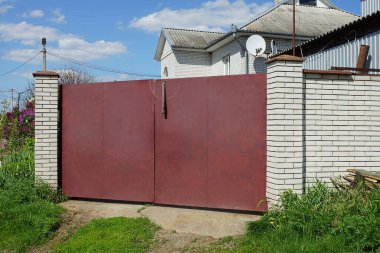 Kapalı bir kırmızı demir kapı ve sokakta yeşil çimenler olan beyaz bir tuğla duvar.