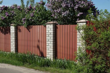 Kırsal bir sokakta yeşil bitki örtüsü ve çimlerle kaplanmış kırmızı metal ve beyaz tuğlalardan yapılmış bir çitin parçası.