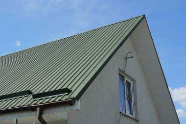 Mavi gökyüzüne karşı yeşil metal çatının altında penceresi olan özel bir evin gri tavan arası.