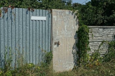 Paslanmış eski gri metal bir kapı ve kırsal bir sokakta yeşil çimlerle kaplanmış çitin bir kısmı.