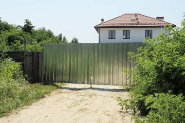 Kırsal bir sokakta yeşil bitki örtüsüyle kaplı ahşap bir çitin üzerindeki büyük gri metal bir kapı.