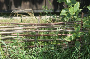 Kırsal bir sokakta yeşil çimen ve bitki örtüsüyle kaplanmış ahşap dallardan yapılmış dekoratif gri kahverengi bir çitin parçası.