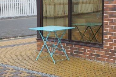 one blue metal table stands on a brown sidewalk outside against a brick wall with a large window