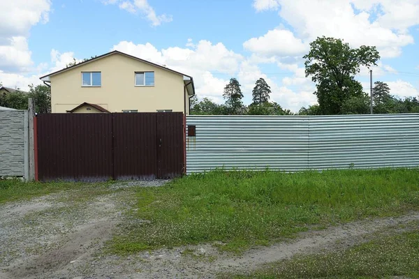 brown iron gate and door on a gray long fence wall in green grass on a rural street in front of a private house against the sky
