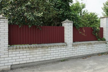 part of the wall of a fence made of red metal and white bricks overgrown with green vegetation  on a rural street