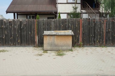 one brown wooden dog house stands on a gray sidewalk against a black fence wall