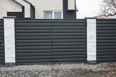 closed black metal gate on a fence wall made of iron and white concrete pillars on a rural street