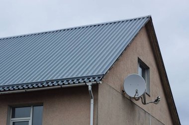 Gri gökyüzüne karşı yeşil metal bir çatı altında penceresi ve uydu anteni olan kahverengi tavan arası.
