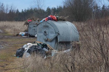 Bir gri metal çöp tenekesi sokaktaki kuru çimlerin üzerinde duruyor.