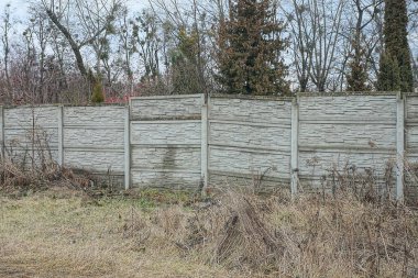 Kırsal bir sokakta, kuru çimlerin üzerinde uzun, gri beton bir duvar.