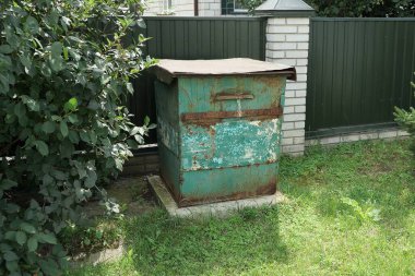 Eski paslı, kapalı metal bir çöp tenekesi yeşil çimlerde ve kırsal bir sokakta, demir çitlerin yanında duruyor.