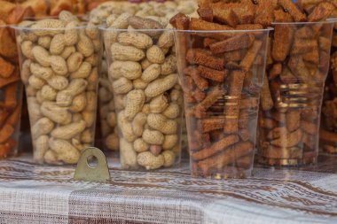 Masadaki beyaz plastik bardaklarda gri kabuklu fıstıklar ve kahverengi kuru krakerler.