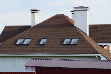 Pencereleri ve gri gökyüzüne karşı beyaz beton bacaları olan özel bir evin kahverengi kiremitli çatısı.