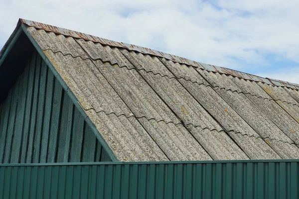青い空と白い雲に対する緑の金属製のフェンスの後ろの田舎の家の灰色のスレート屋根 — ストック写真