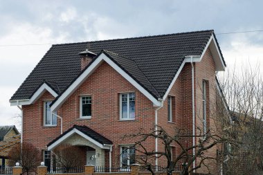 Gri gökyüzüne karşı siyah kiremit çatı altında pencereleri olan büyük kahverengi tuğlalı bir ev.