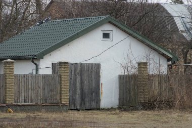 Kırsal bir sokakta gri bir çitin arkasında yeşil kiremitli çatının altında küçük bir penceresi olan beyaz bir ev.