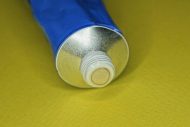 Sarı bir masanın üzerinde kahverengi yapıştırıcılı açık, büyük, beyaz, gri bir tüp var.